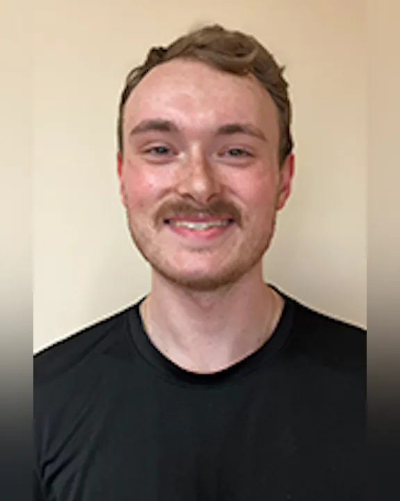 Logan Grevelding with short hair and a mustache, wearing a black shirt, smiling against a plain background.