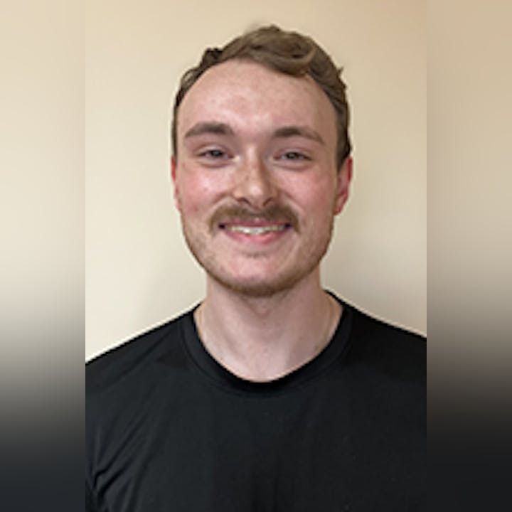 Logan Grevelding with short hair and a mustache, wearing a black shirt, smiling against a plain background.