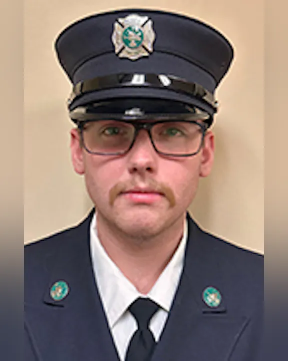 Colin Decker in a firefighter uniform and hat, wearing glasses, with a mustache, against a plain background.