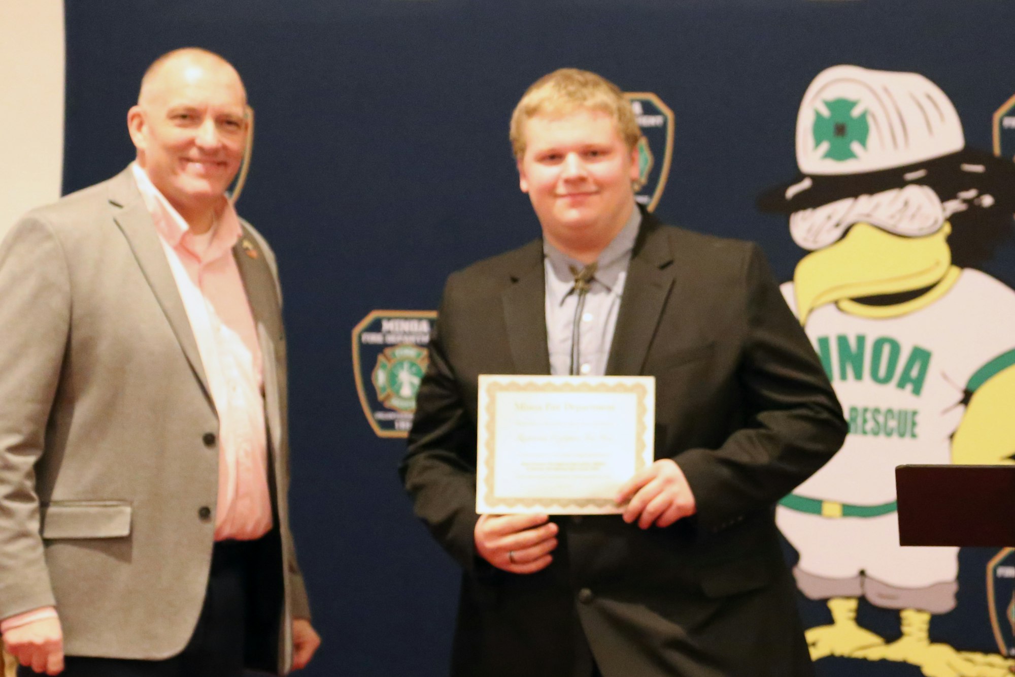 Two people posing; one holds a certificate. A cartoon mascot character in the background.