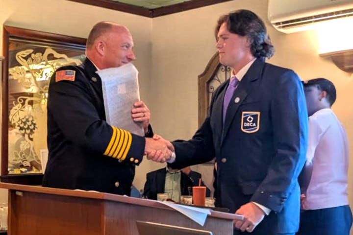 Chief Grevelding and Firefighter Reynolds are shaking hands, the Chief in a uniform with an American flag patch, Reynolds in a blazer with a DECA badge.