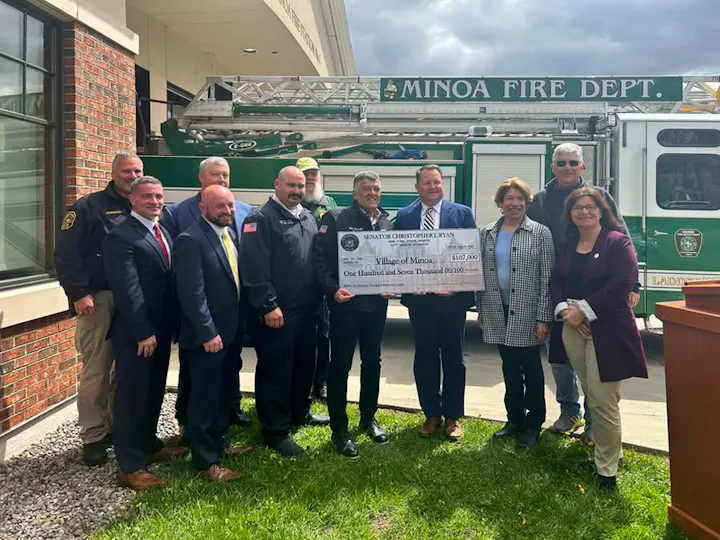 A group of officials presents a large check for $107,000 to the Village of Minoa, with a fire truck in the background.