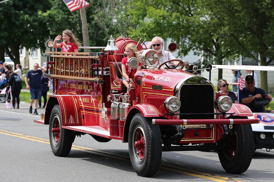 Vintage red fire truck in the parade with people aboard, waving to onlookers, American flag in the background.
