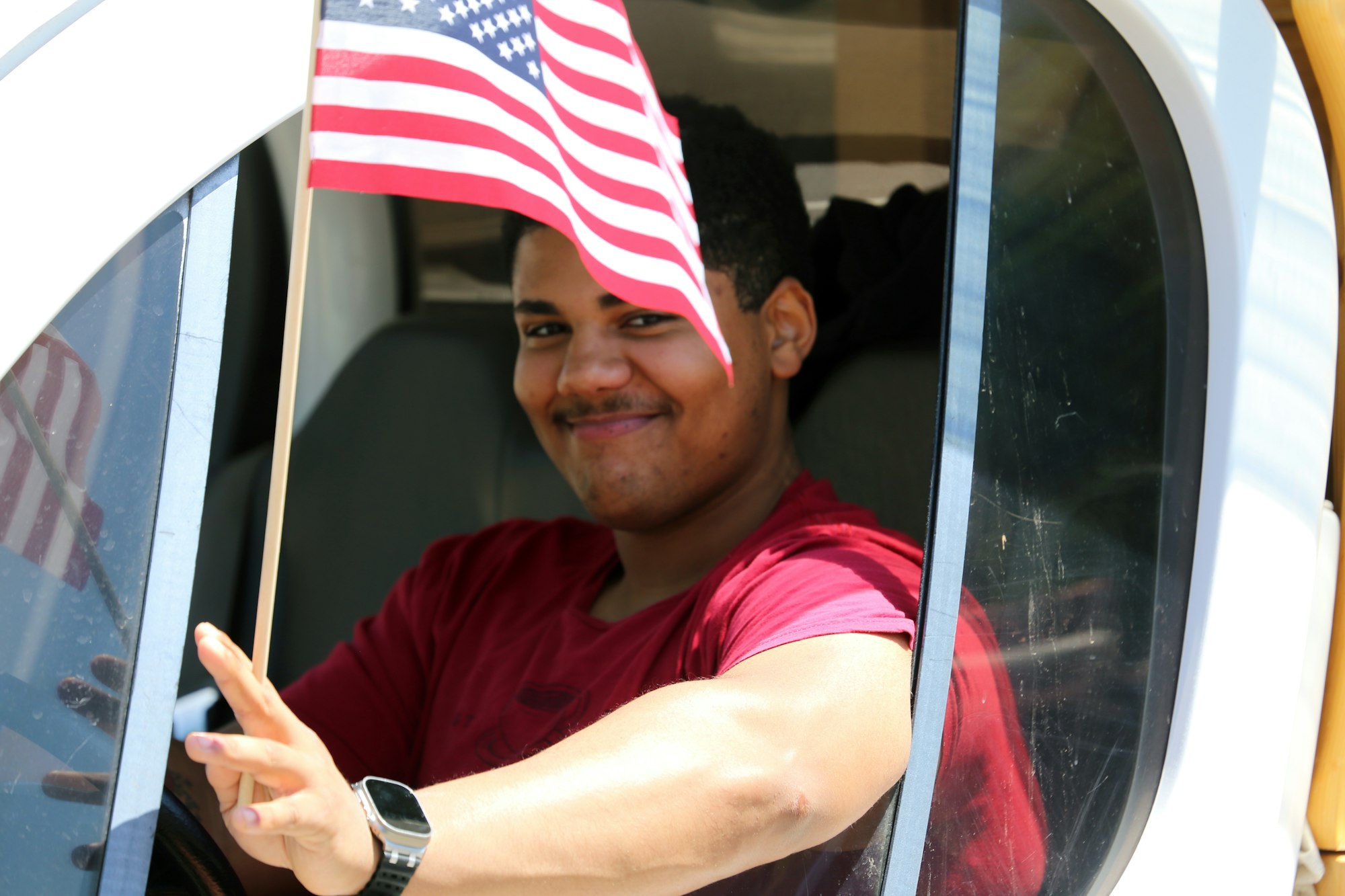 Person in a vehicle, smiling, wearing a red shirt and watch, holding a small American flag.