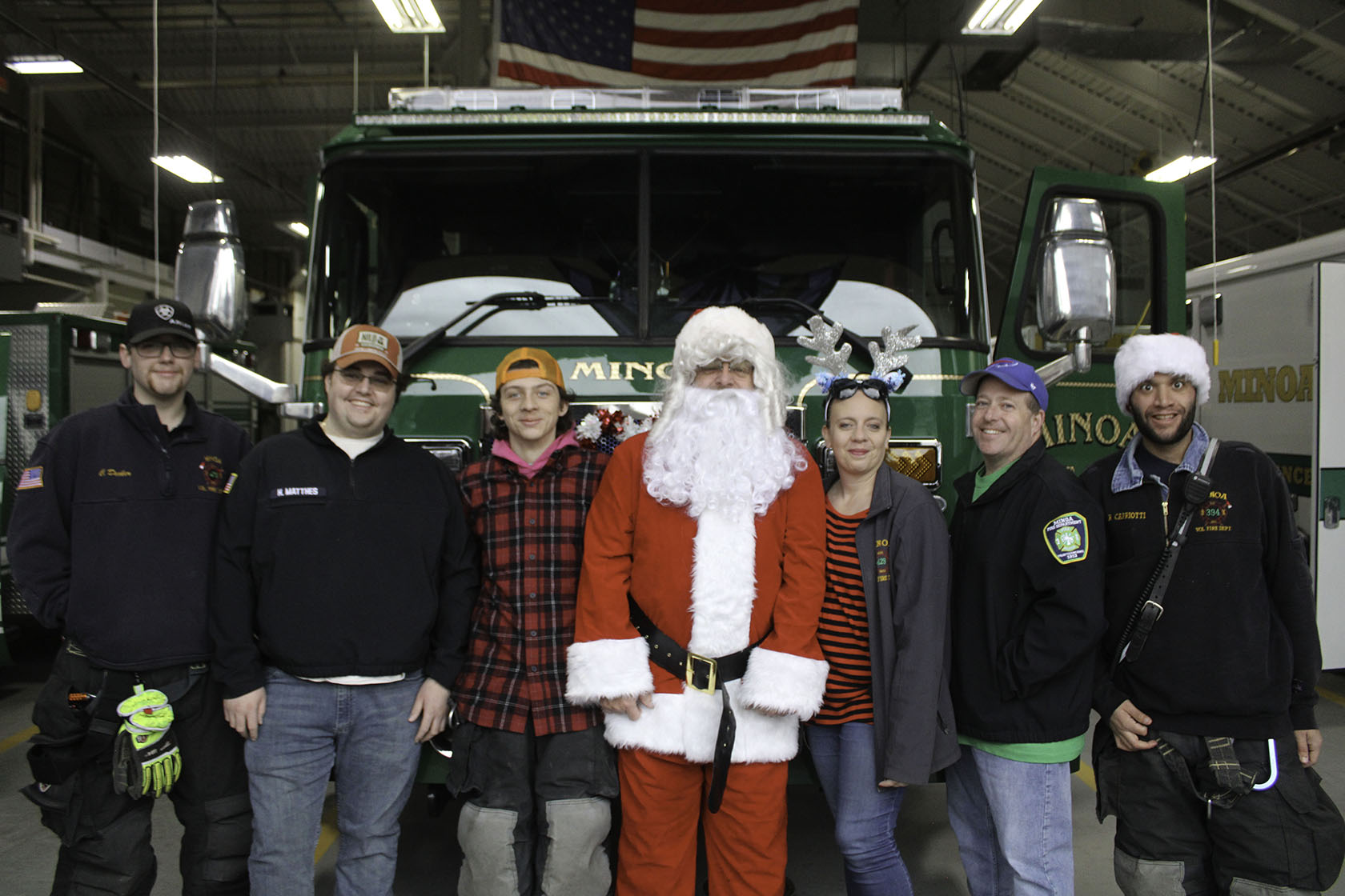 Group of people, including Santa Claus, posing in a fire station with a fire truck and American flag in the background.