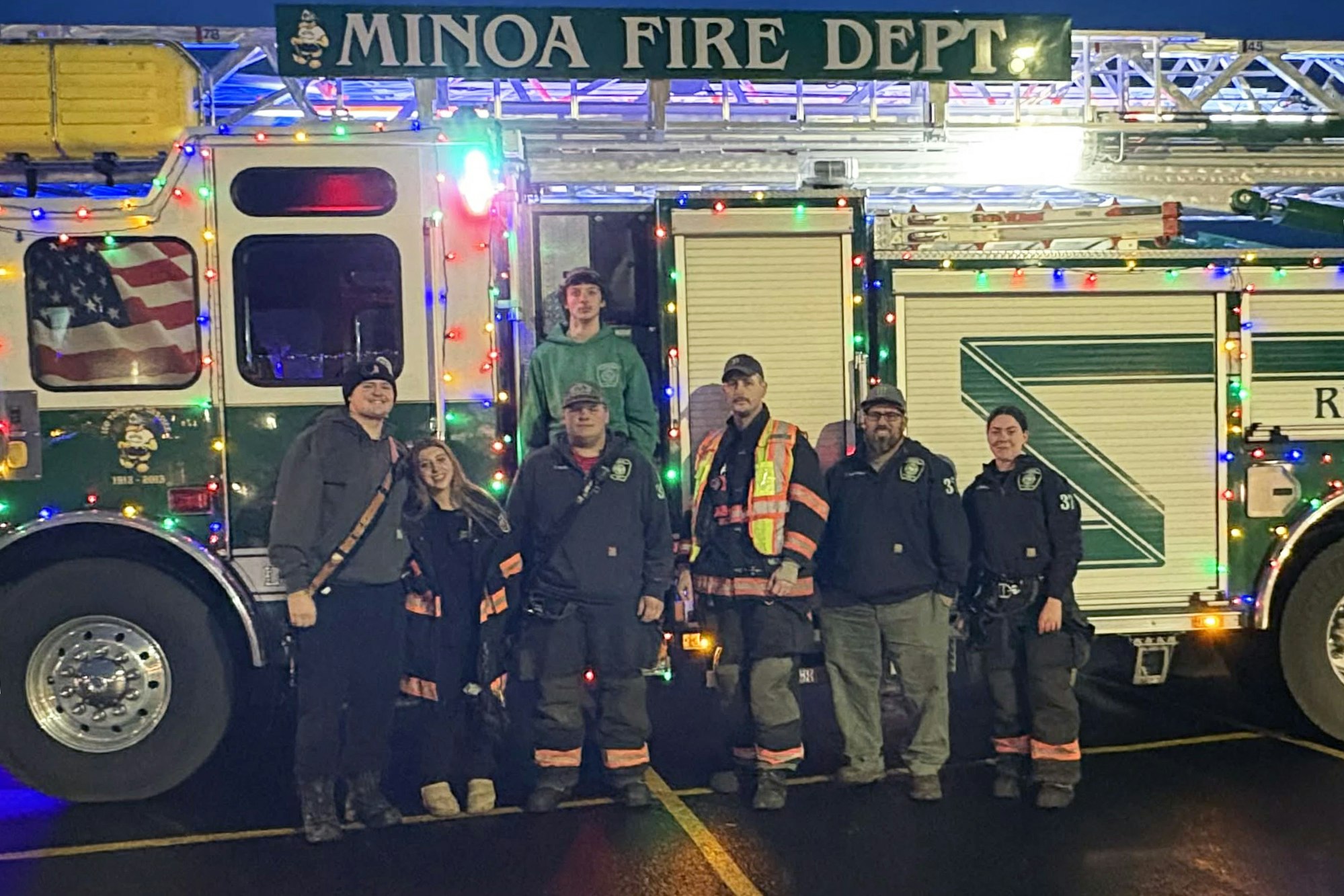 A group of firefighters stands in front of a decorated fire truck, showcasing holiday lights and their uniforms.