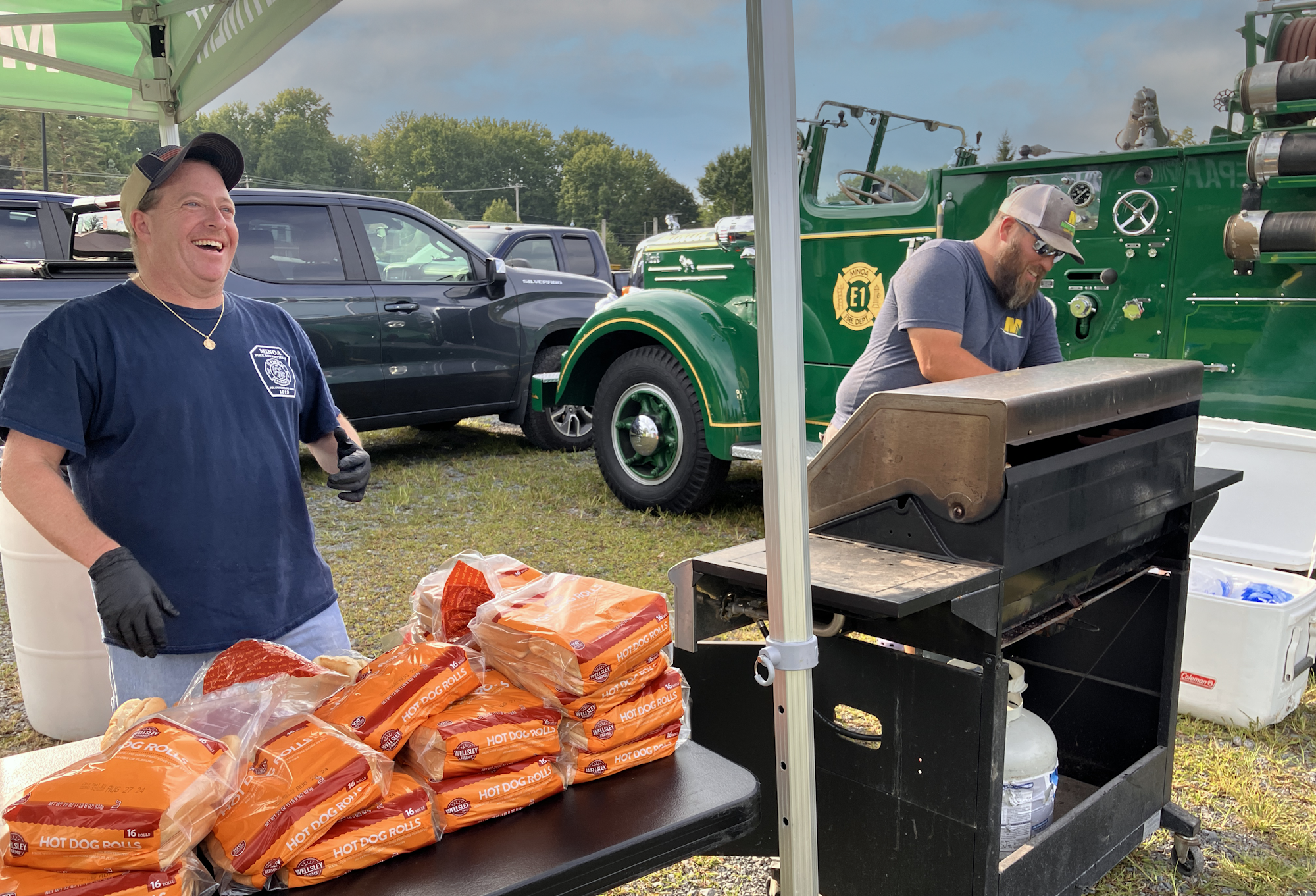 Two men near a BBQ grill with hot dog rolls, a vintage fire truck in the background. Both men are having a good time.