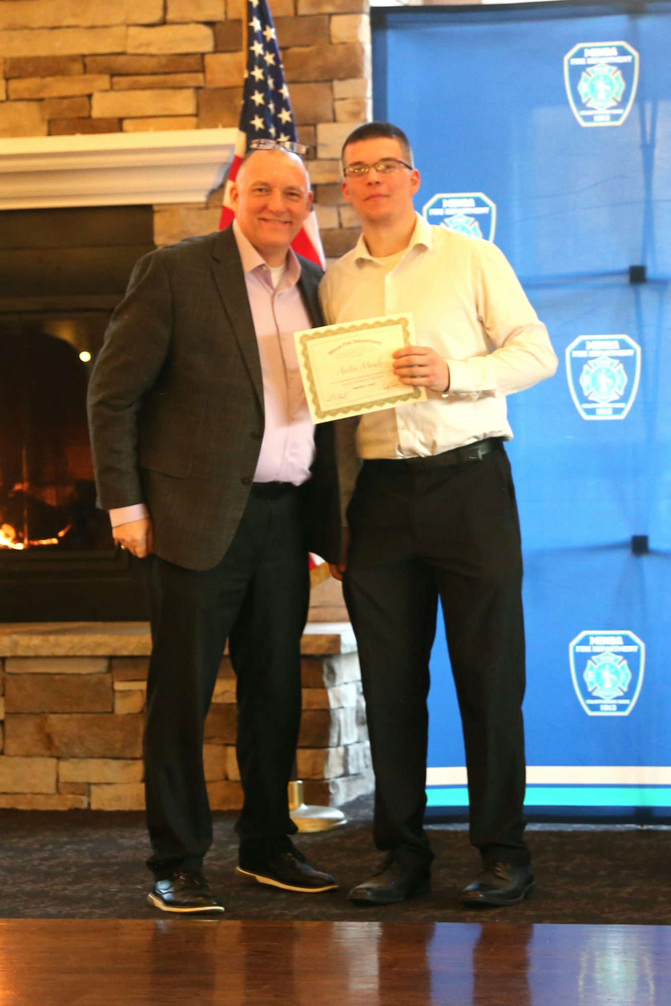 A formal event where one individual receives a certificate, posed with another person in front of an American flag and a backdrop.
