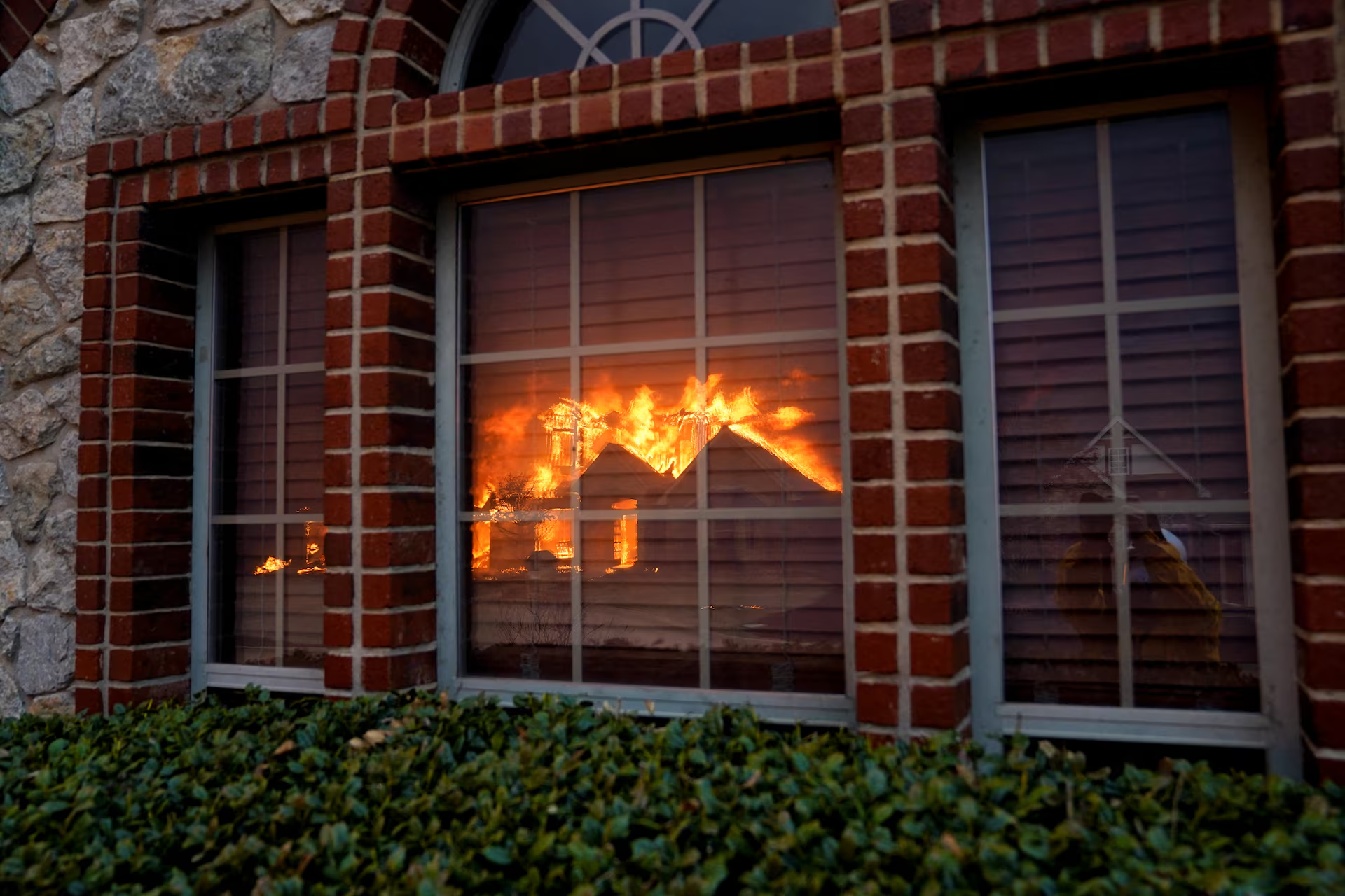 In stillwater, a house fire reflected in the window of a brick building, behind some shrubs.