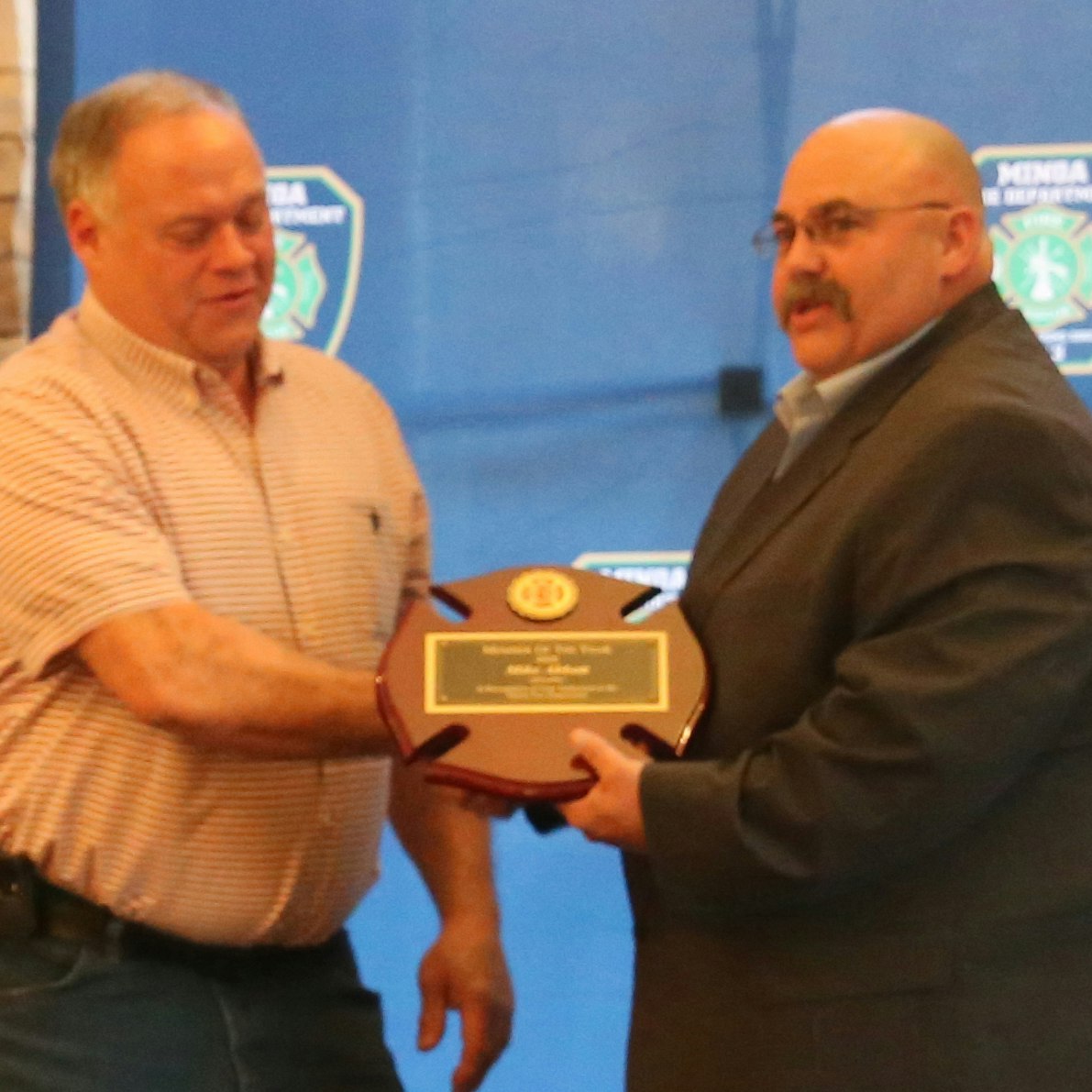 Two men are shaking hands, with one presenting an award plaque to the other at an official event.