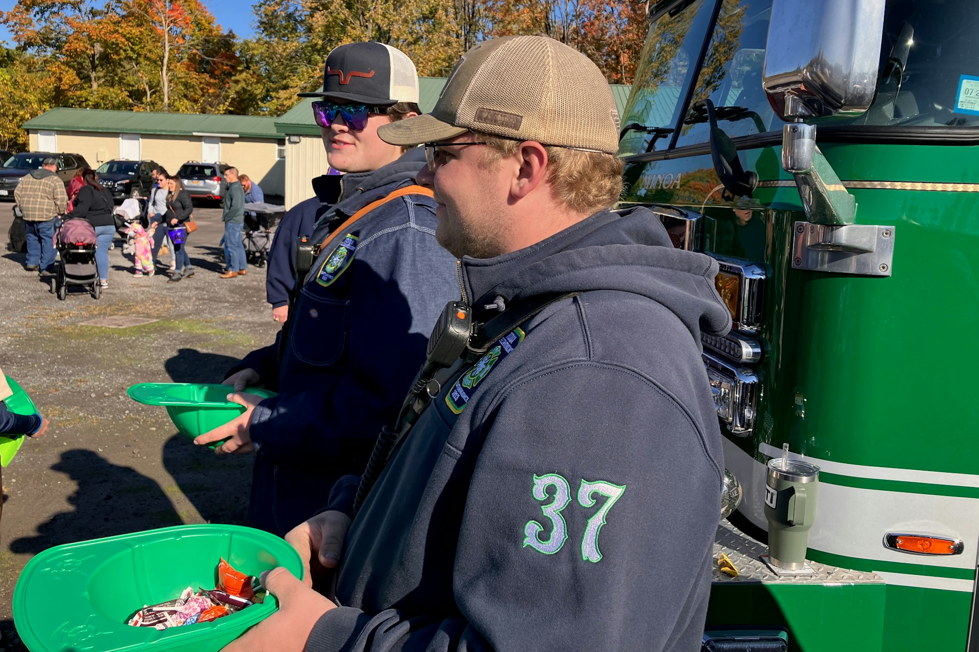 Two men holding green plastic replicas of firefighter helmets with candy, one wearing a jacket with "37" on it, by a shiny green truck, with people and fall trees in the background.