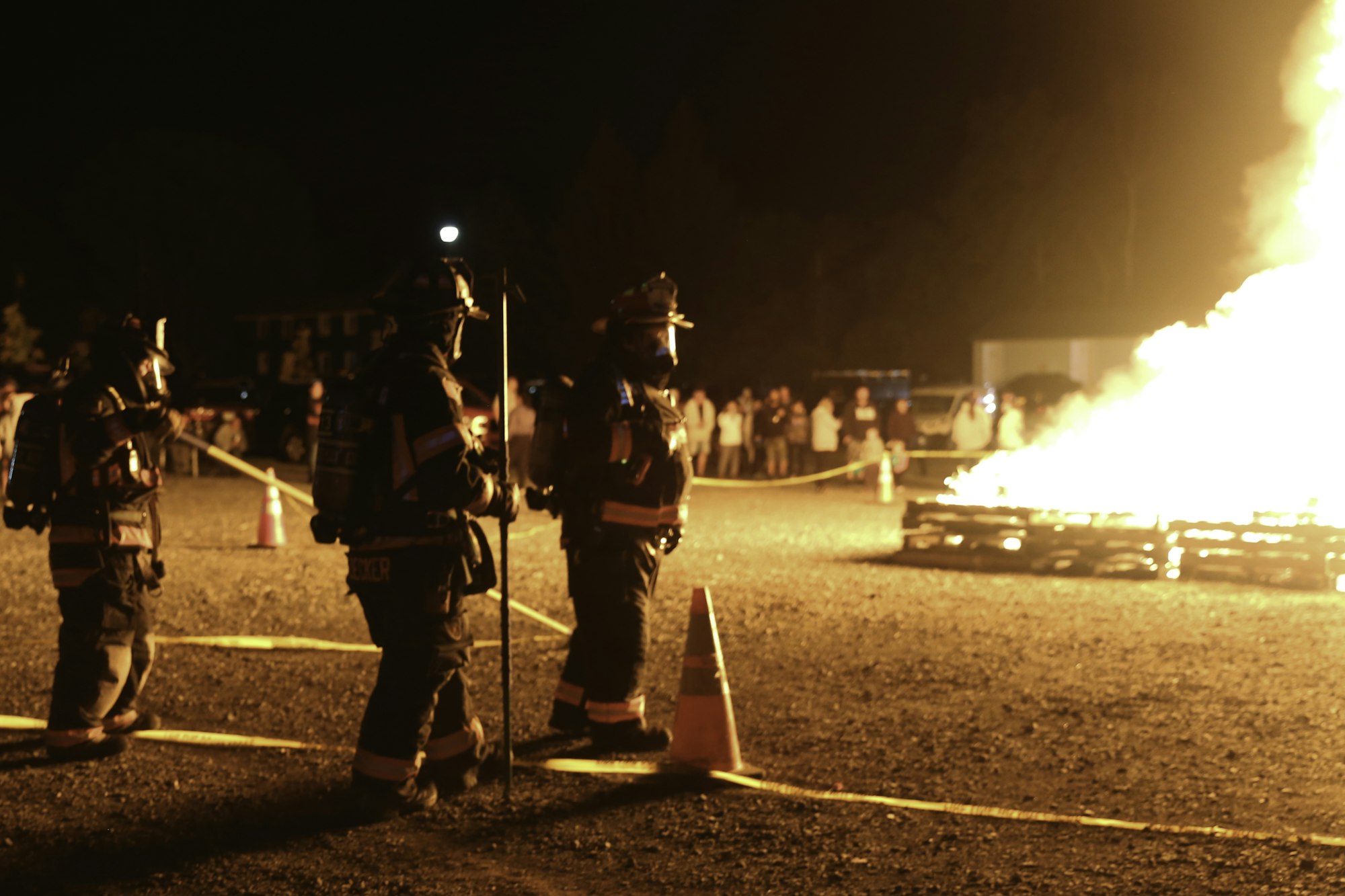 Firefighters near a large fire at night, with onlookers in the background.