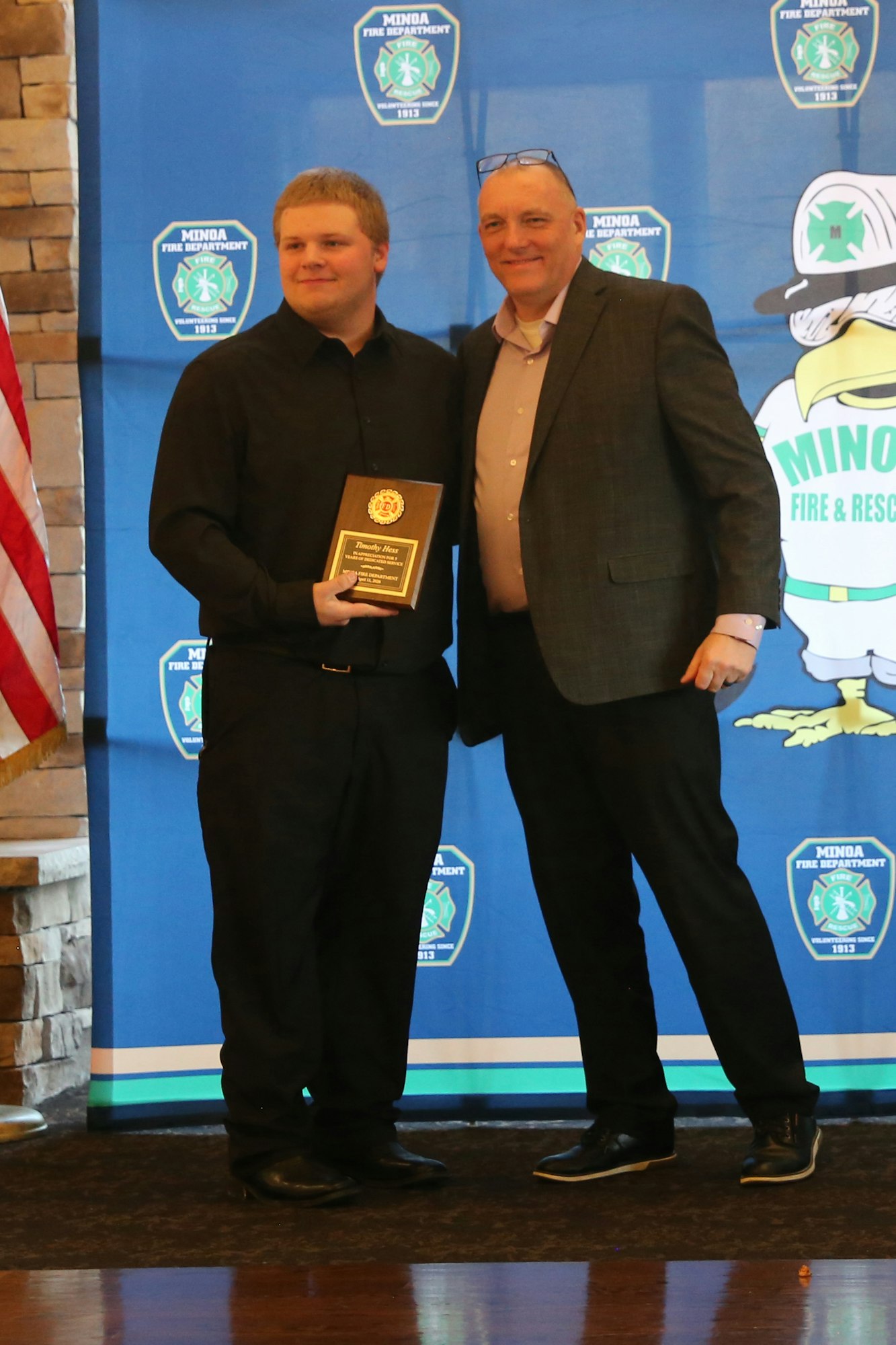 A young man receives an award from another man during a ceremony, with a backdrop featuring fire department logos.