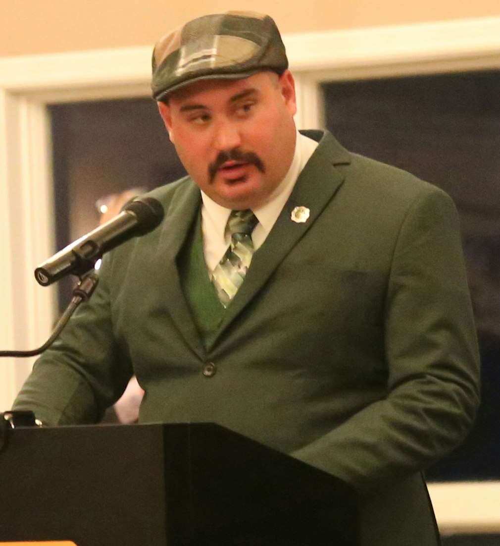 A man in a green suit and cap speaks at a podium with a microphone, likely at a formal event.