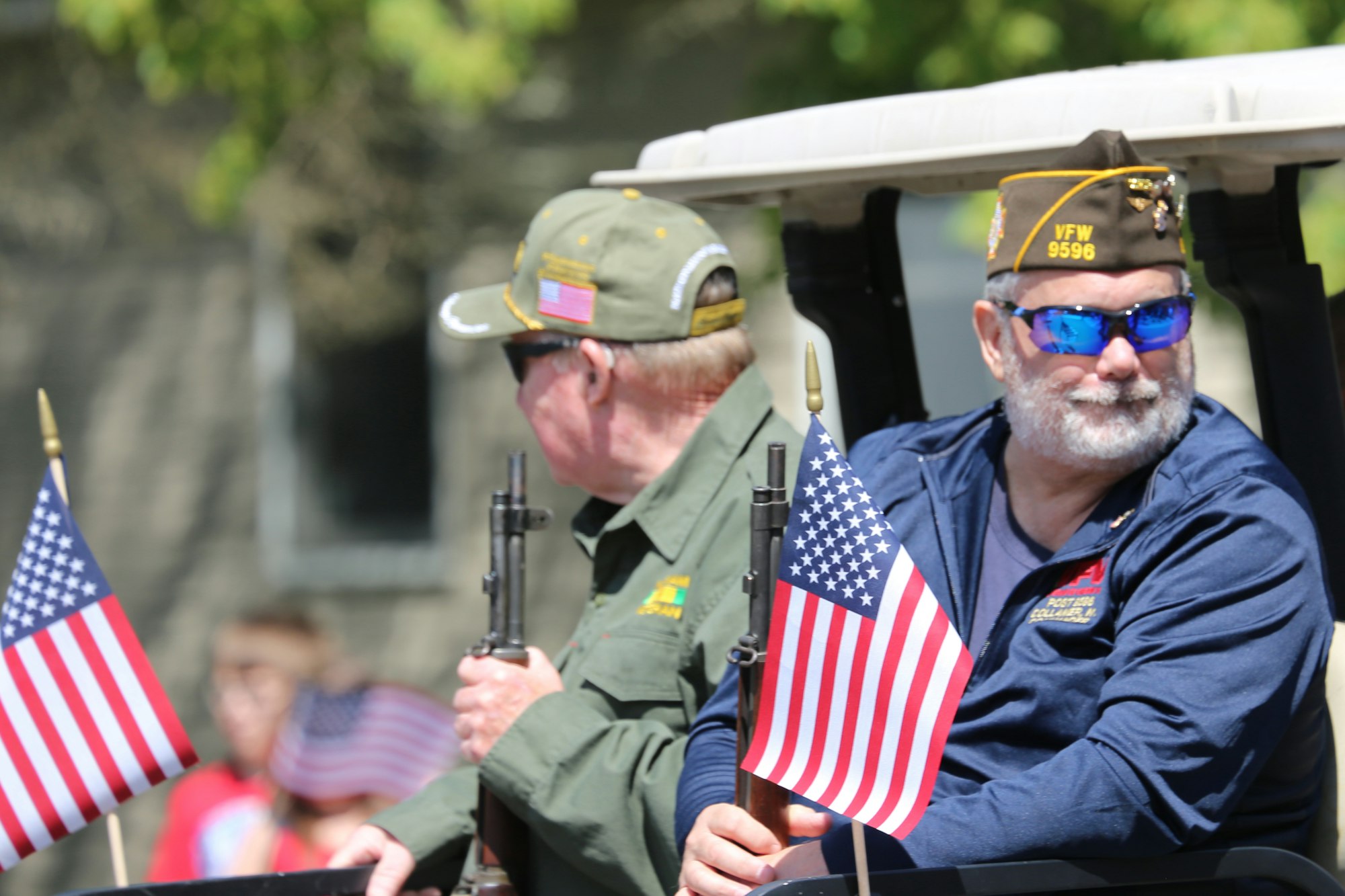 Two veterans in a vehicle holding rifles and American flags during a parade.