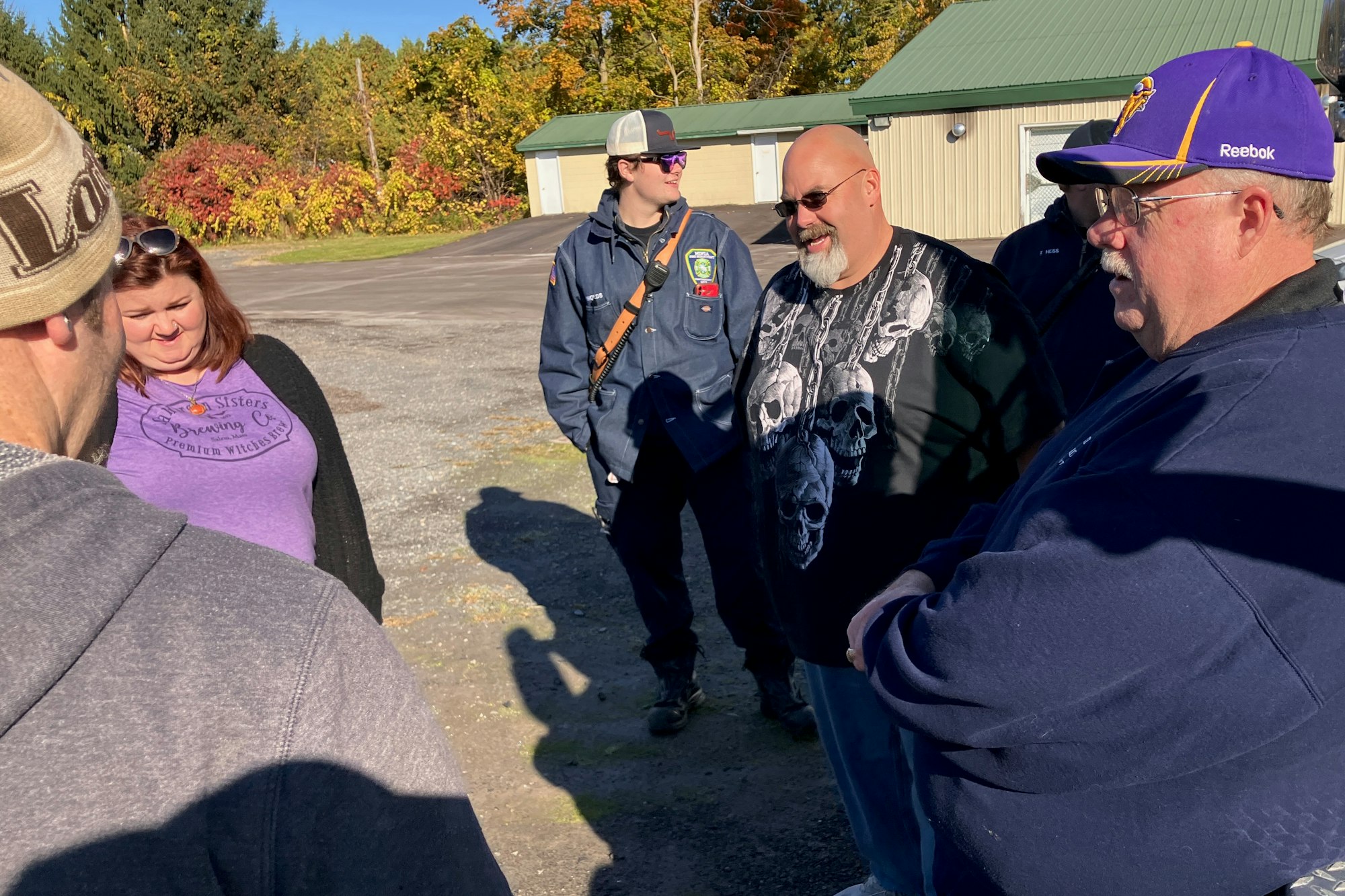 A group of people connected to the Minoa Fire Department standing in a parking area, smiling and engaging in a conversation, with autumn trees in the background.