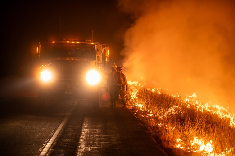 Two people in firefighting gear walk beside a large fire truck near a wildland blaze at night.