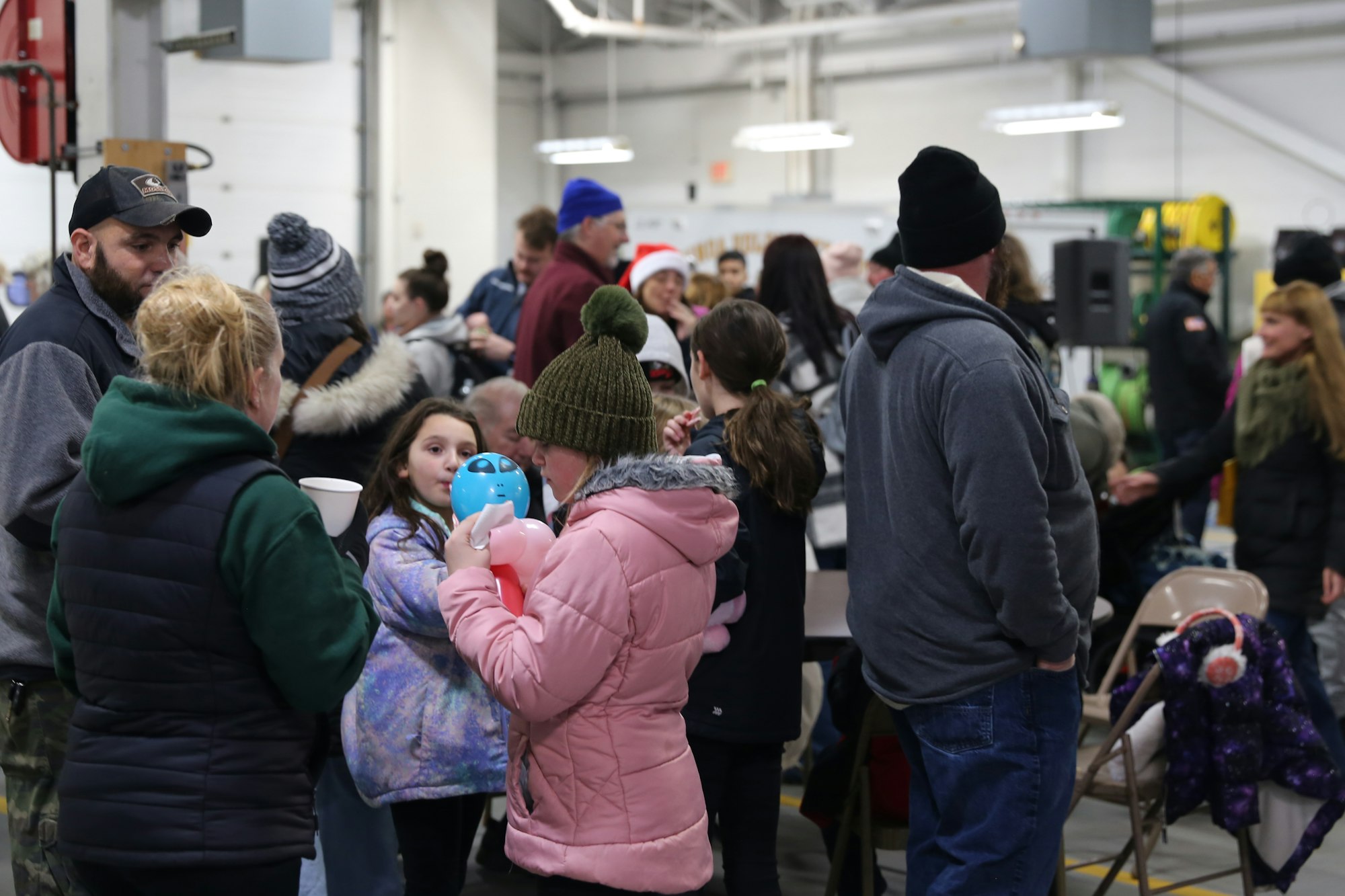 A group of people gathered indoors, some in winter clothing, one child holding balloons.