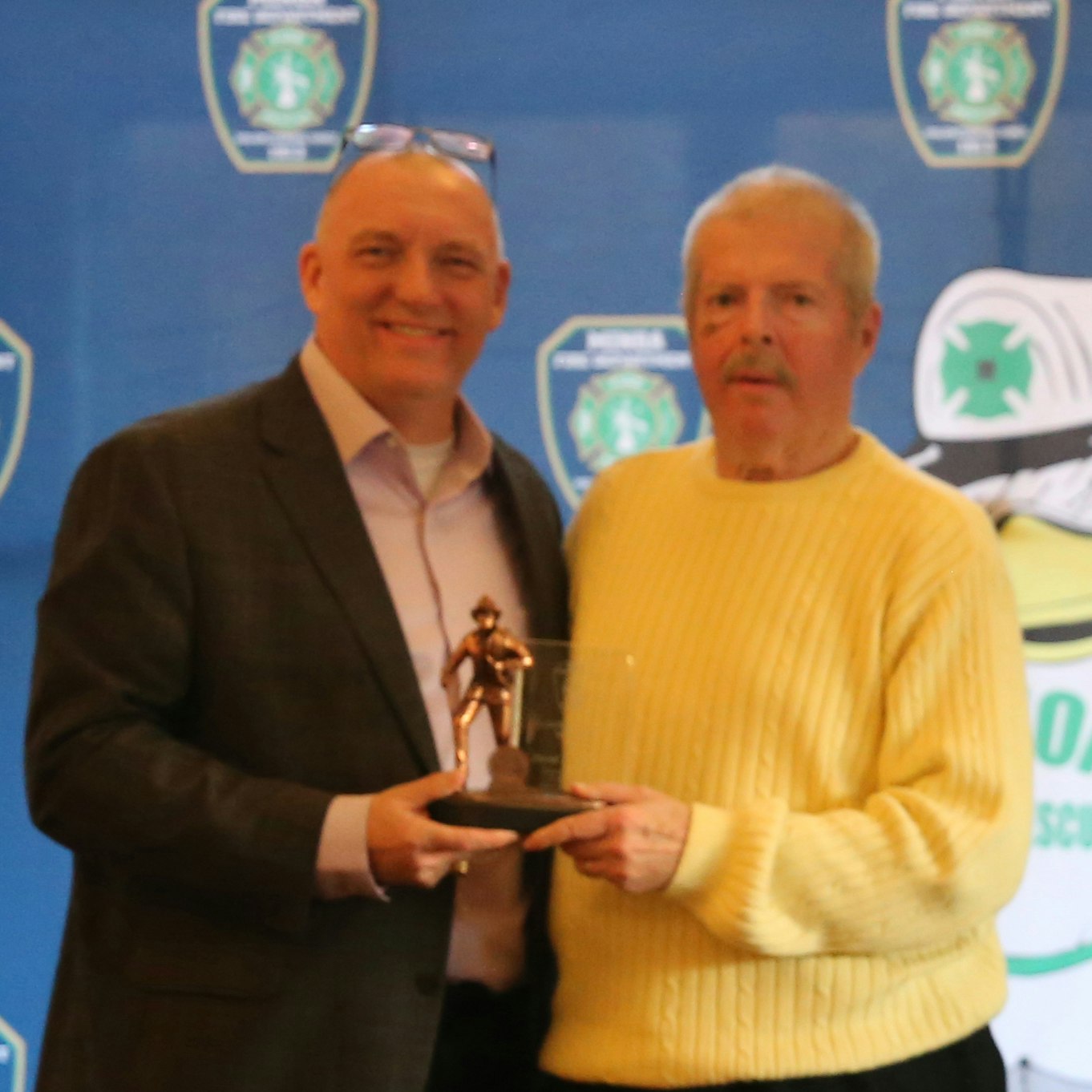 Two men are smiling and posing with an award or trophy at an event, with a blue background featuring logos.