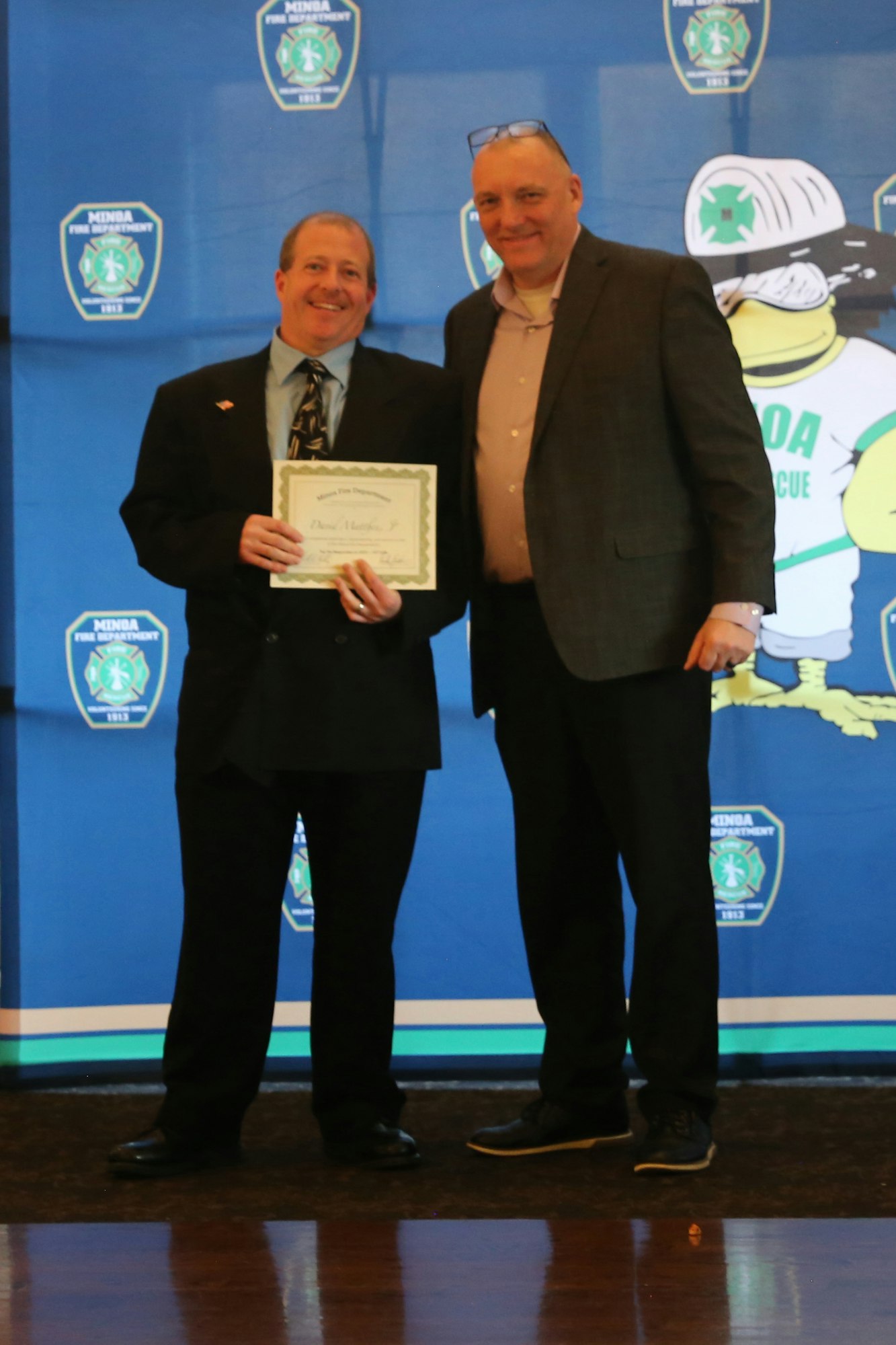 Two men stand together, one holding a certificate, against a backdrop featuring the Minda Fire Department logo.