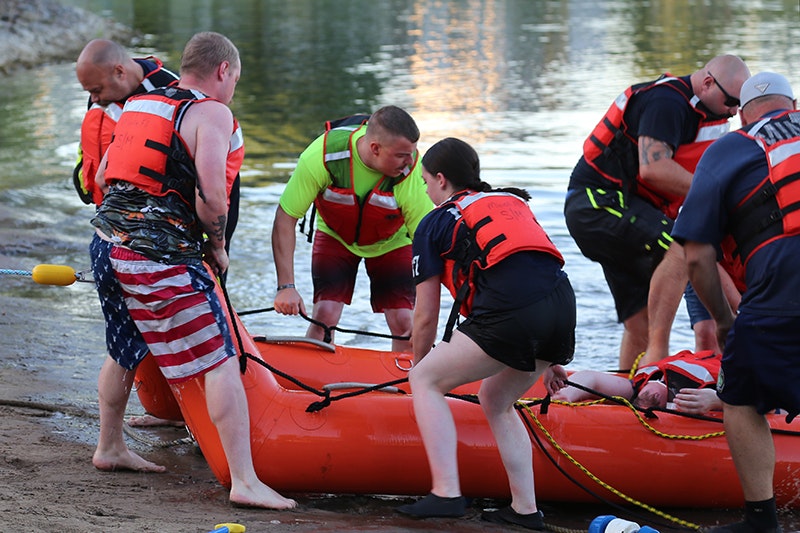 A group of firefighters in life vests work together to move an inflatable orange canoe with a victim on a shoreline.