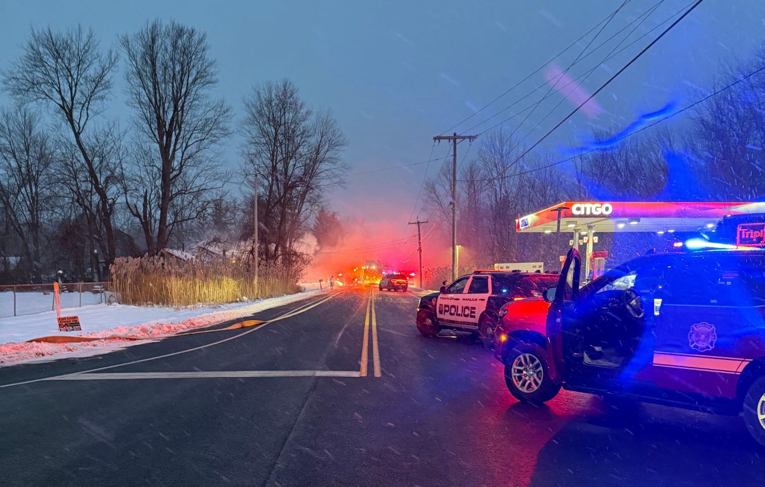 Emergency scene with police and fire response vehicles on a snowy road, near a gas station. Visibility is low due to weather.