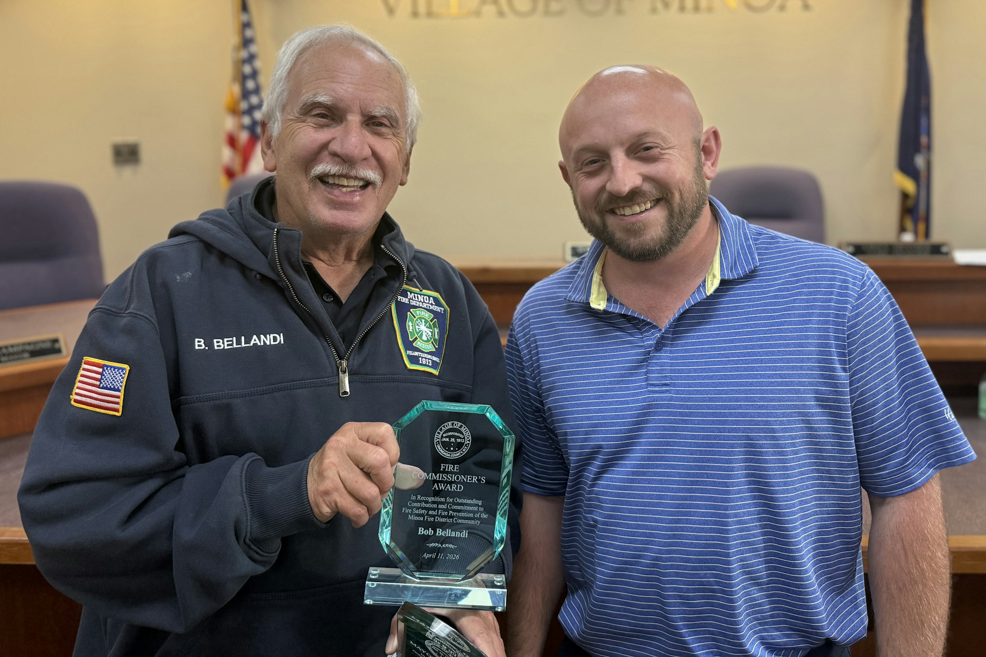 Bob Bellandi and Fire Commissioner Bobby Schepp are smiling together, one holding a Fire Commissioner's Award plaque, in the village board's meeting room.