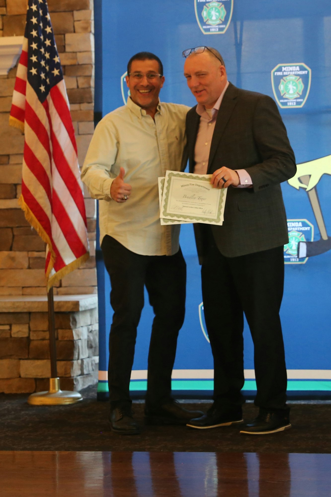 Two men are smiling and posing with a certificate at an event. An American flag is visible in the background.