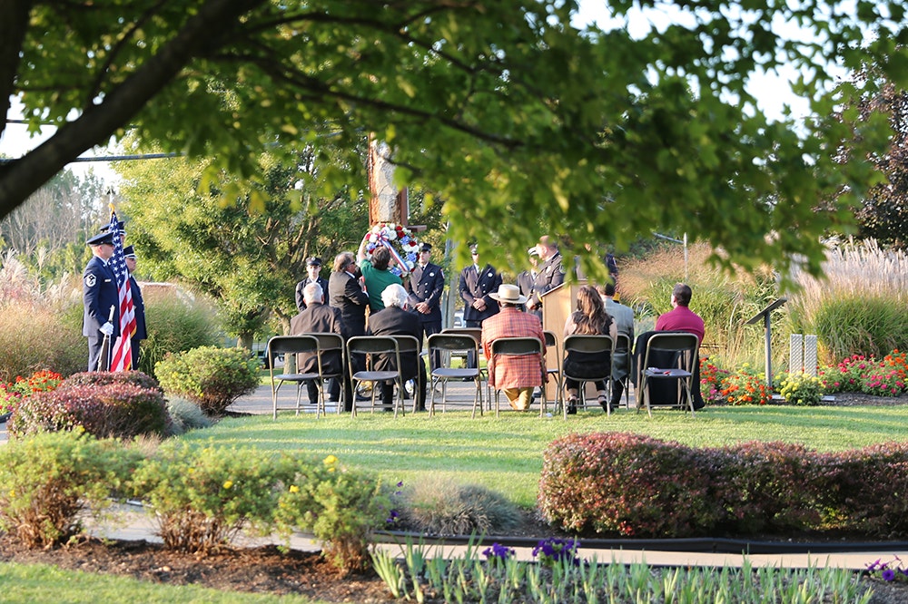 A group of people attend hte 9-11 memorial commemoration in an outdoor ceremony with uniformed personnel, a flag presentation, and a wreath in a garden setting.