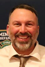 A smiling Todd Durantini with a beard, wearing a shirt and tie, standing in front of a badge-like background.