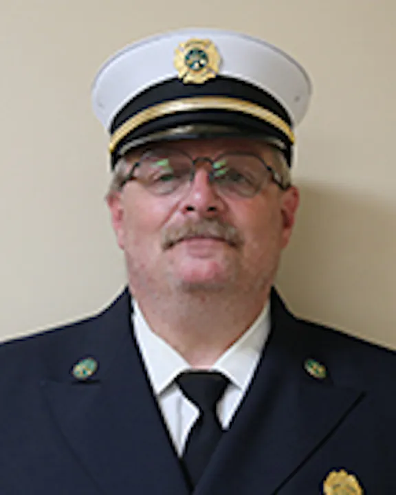 Dave Hess in a white firefighter's uniform cap with a badge, wearing glasses, and a mustache.