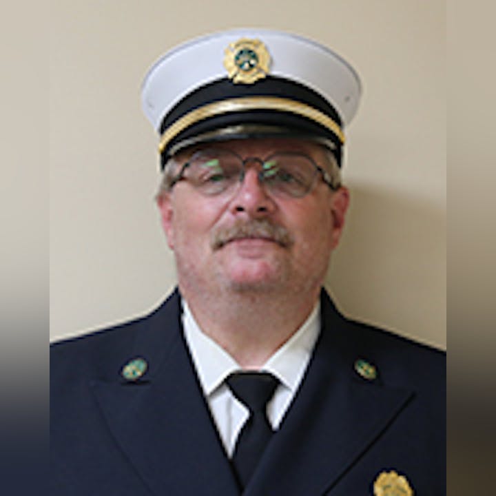 Dave Hess in a white firefighter's uniform cap with a badge, wearing glasses, and a mustache.