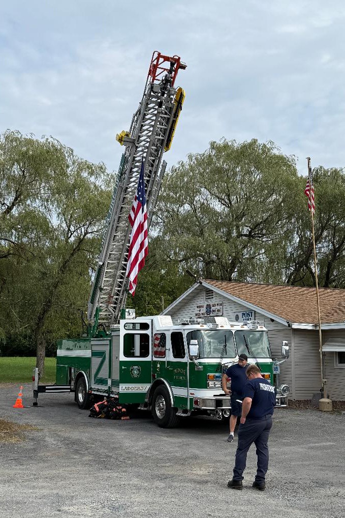 A green fire truck with an extended ladder, an American flag, and two people nearby outside a building.