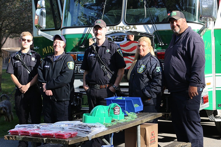Members of the Minoa Fire Department in uniforms standing in front of a Minoa fire truck with tables, helmets, and tools in foreground.