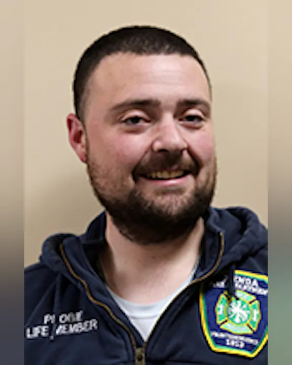 A smiling Matt Rushlo wearing a dark jacket with a badge, against a neutral background.