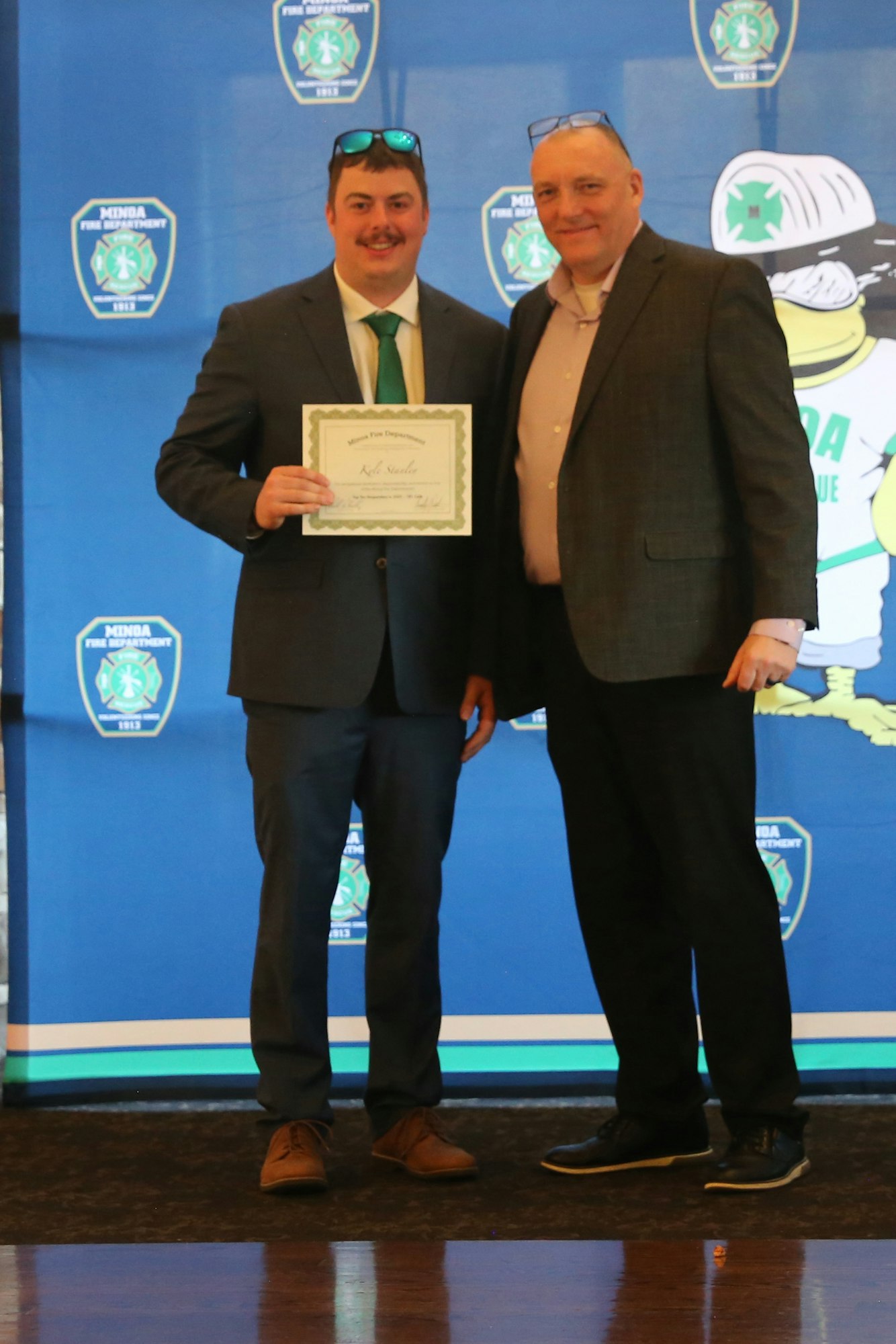 Two men stand together, one holding a certificate, in front of a blue backdrop featuring a fire department logo.