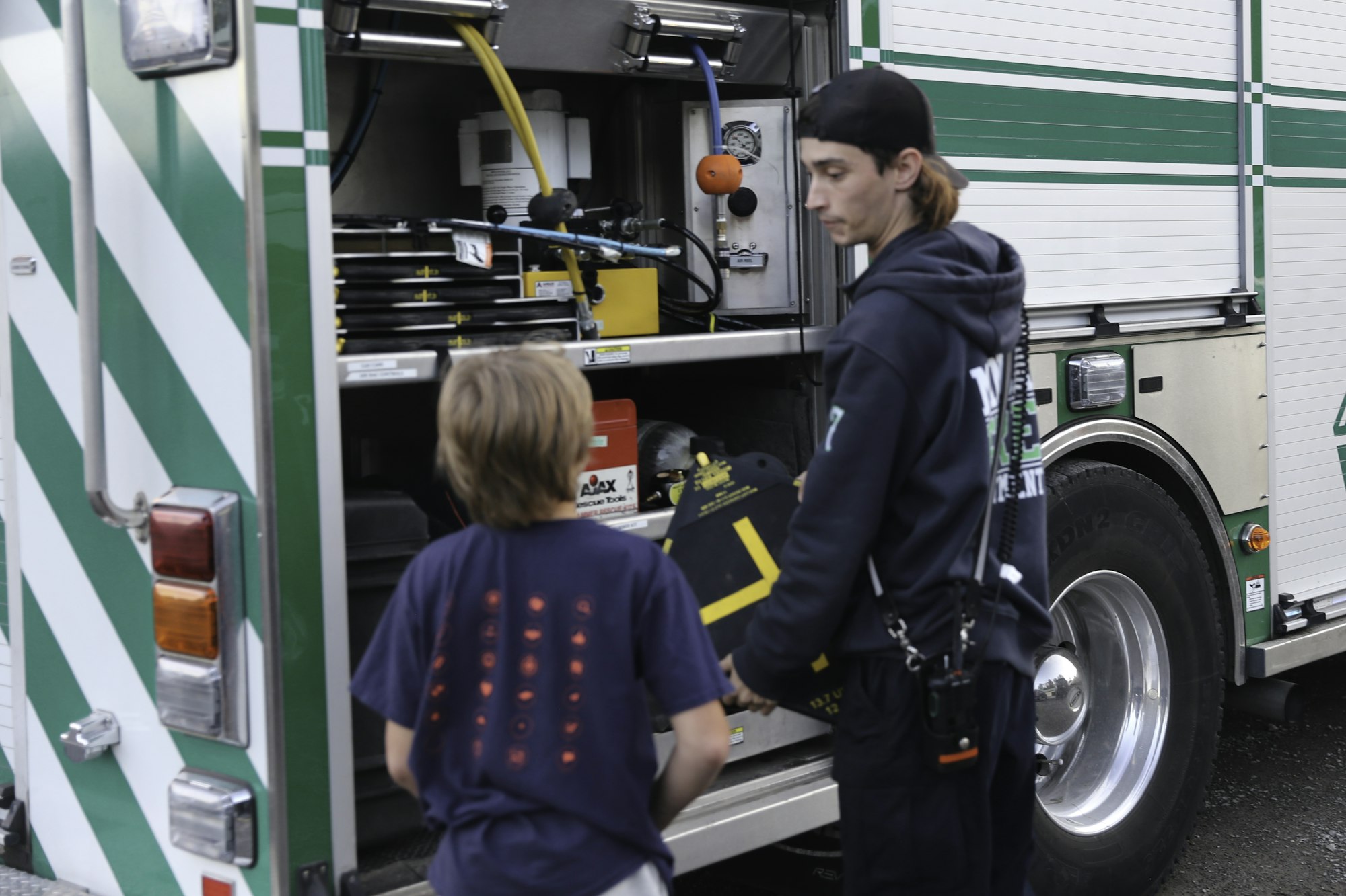 A person in a hoodie and cap showing equipment inside a firetruck to a curious child.