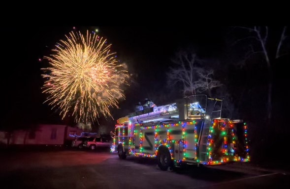 The Minoa Ladder Tuck decorated with colorful lights is parked at night with fireworks exploding in the sky above.