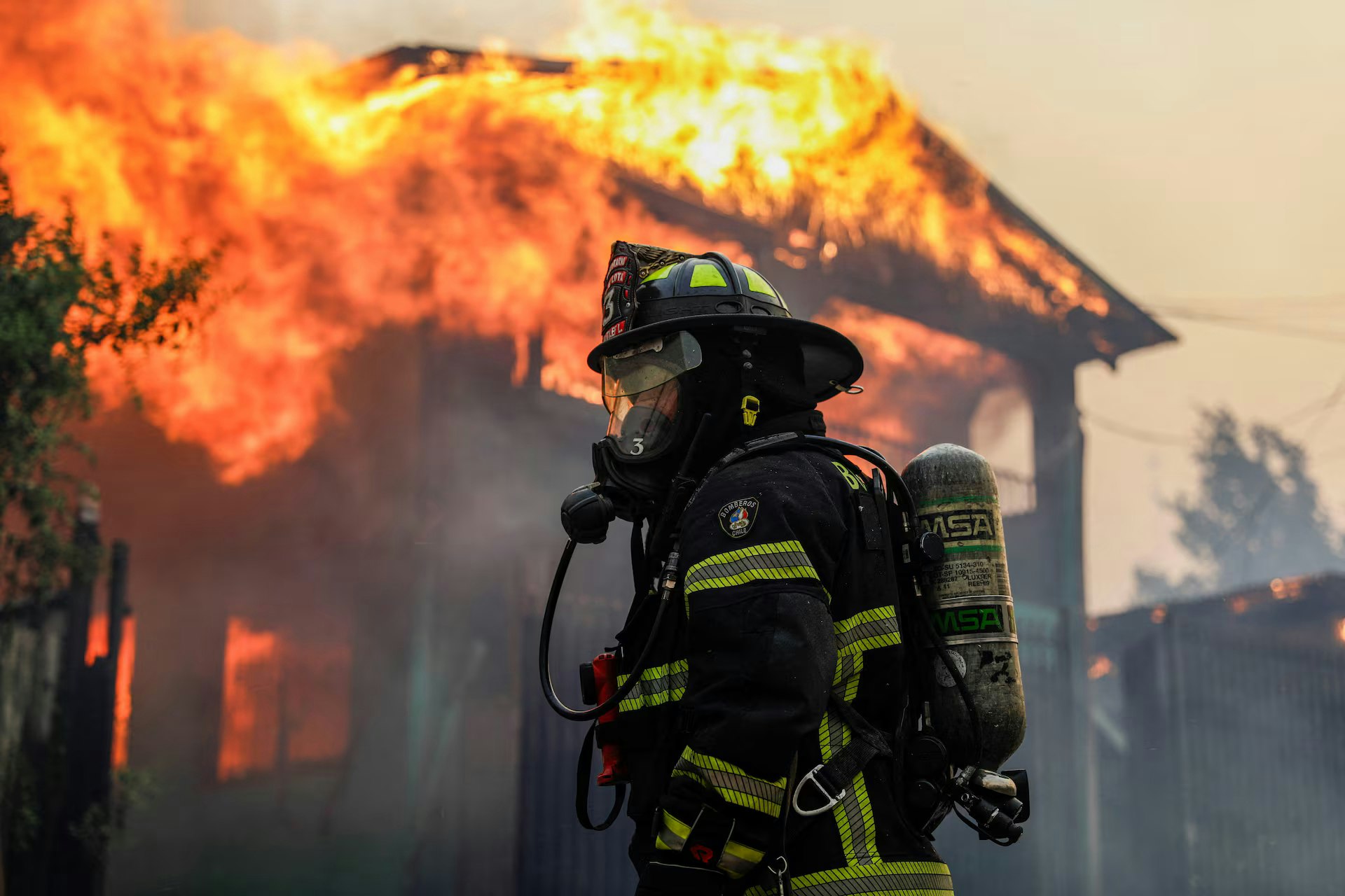 A firefighter stands ready as a building burns fiercely in the background, surrounded by smoke and flames.