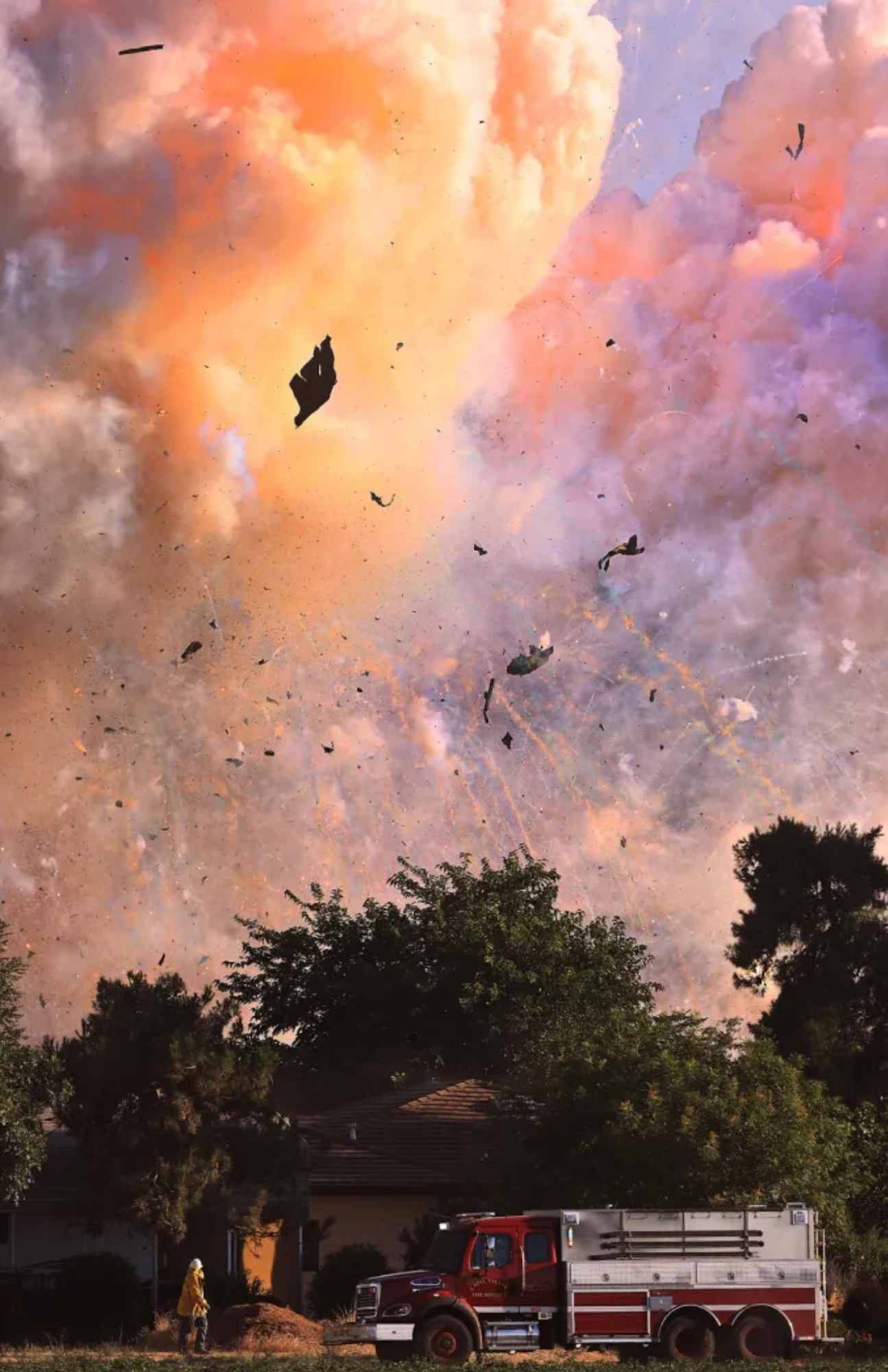 Large explosion with debris flying, massive smoke cloud, firetruck, and firefighter nearby.