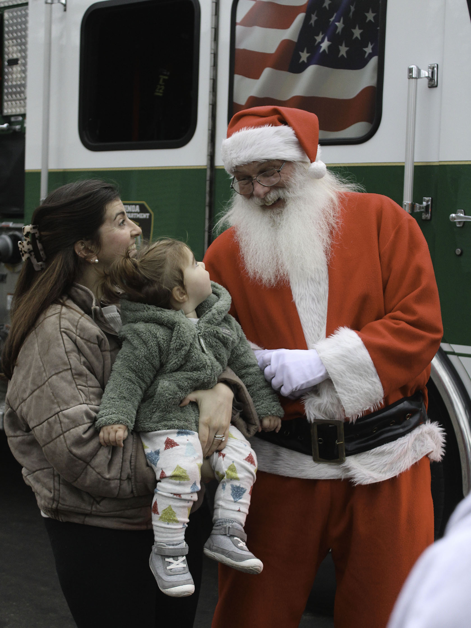 A person dressed as Santa Claus interacts with a smiling woman holding a young child, in front of an ambulance with an American flag.