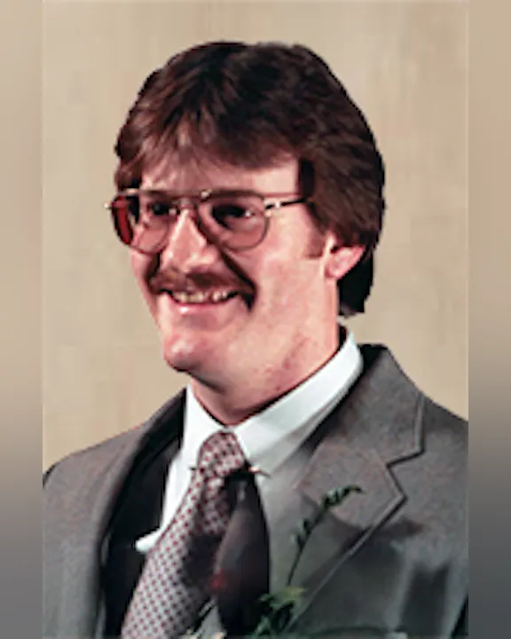 Gary Stoddard in 1984, wearing glasses, a suit, and a tie, smiling against a neutral background.