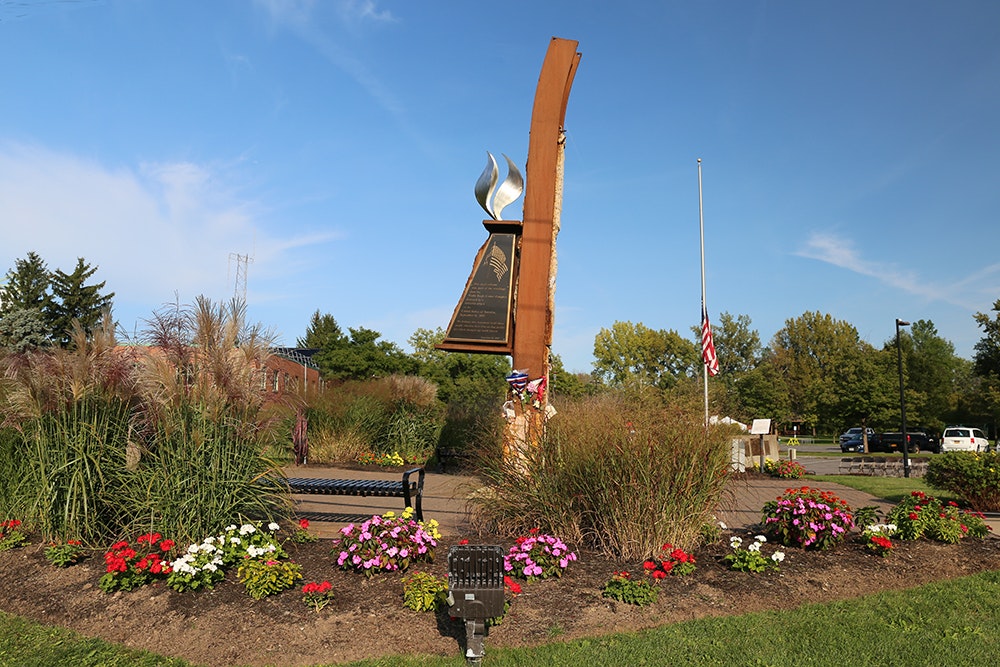 The 9-11 memorial in DeWitt, with a flame motif on a piece of steel from the World Trade Center, surrounded by flowers and flags, under a clear blue sky.