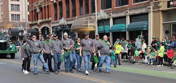 A group of Minoa firefighters in gray shirts walking in a parade, with spectators and buildings in the background.