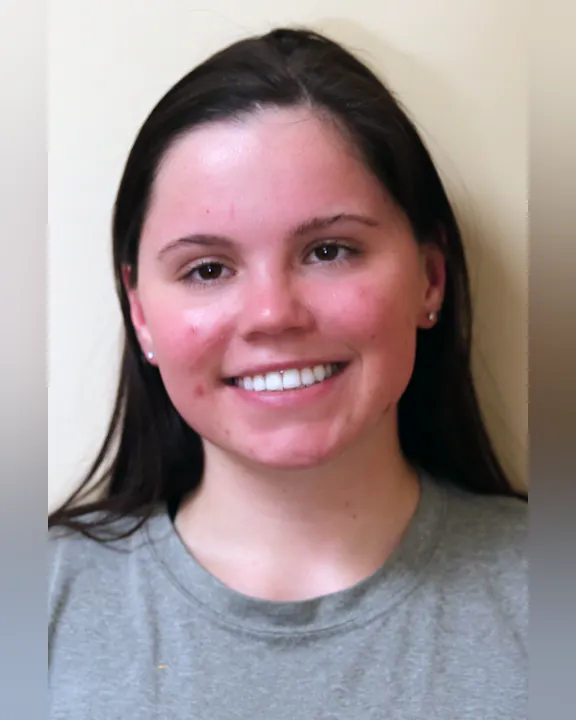 Anna D'Amore with long dark hair is smiling, wearing a gray shirt, against a light-colored background.
