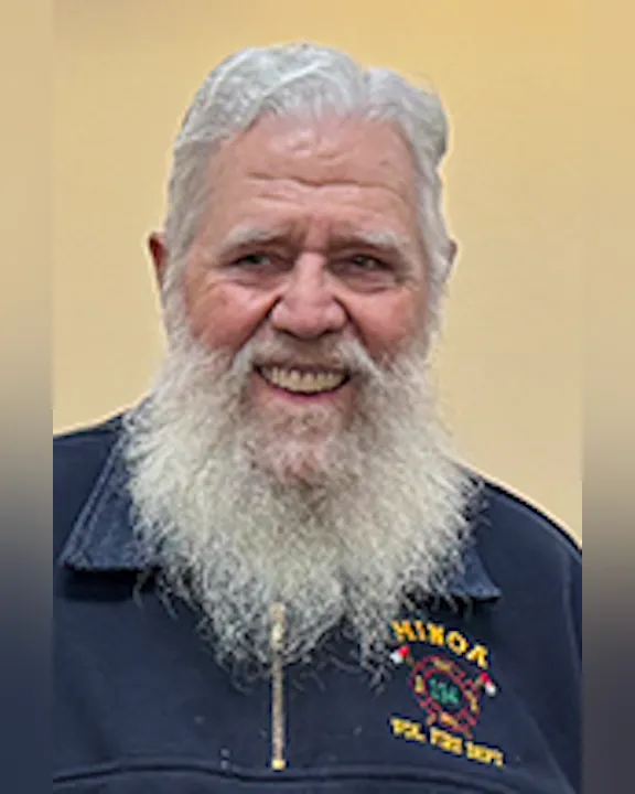 An older man with a long white beard, smiling, wearing a jacket with a fire department logo.