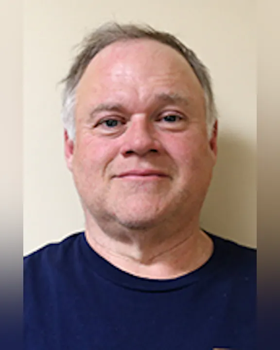 Mike Abbott with short hair wearing a dark blue shirt is standing against a plain light-colored background.