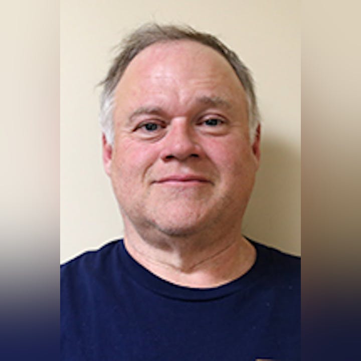 Mike Abbott with short hair wearing a dark blue shirt is standing against a plain light-colored background.