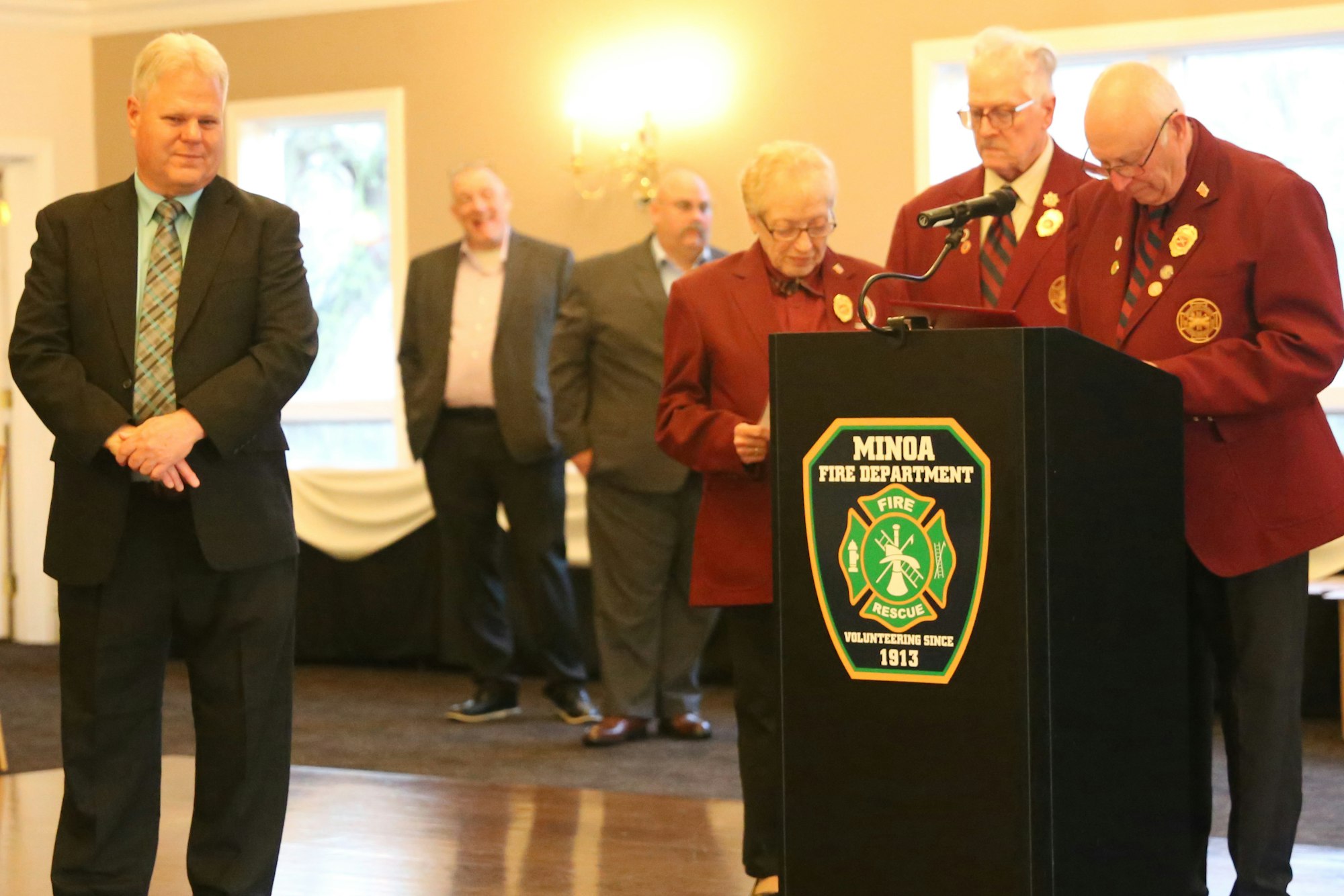 The image shows a group at a ceremonial event for the Minoa Fire Department, featuring speakers and attendees in formal attire.