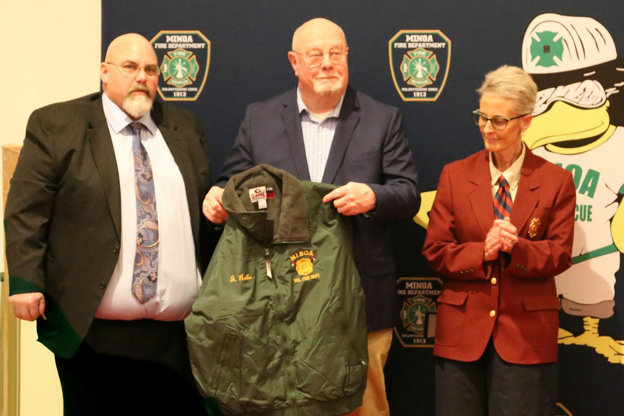 President Decker, Jack Robie (holding his award jacket), and OCVFA Director Tina Cali posing with a Minoa Fire Department jacket in front of a backdrop with fire department logos.
